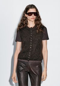 Frau mit langen, welligen Haaren, die eine dunkle Sonnenbrille, eine knöpfbare, flauschige braune Weste über einem Kurzarmhemd und dunkle Lederhosen trägt.