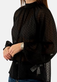 Blusa negra transparente con mangas abullonadas y cuello alto, con un patrón de lunares en relieve. Combinada con un reloj de plata y oro.