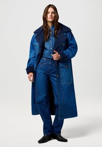 Denim takki, jossa on suuri merensinisestä karvasta tehty kaulus, yhdistettynä denim-paitaan ja leveälahkeisiin housuihin. Kaikki tummansinisessä värissä, jossa on kontrastivärisiä ompeleita ja taskuja.