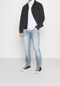 Chaqueta de mezclilla negra, camiseta blanca con pequeños puntos azules, jeans de color azul claro y zapatillas negras con suelas blancas. Atuendo casual, sin patrones ni acentos.