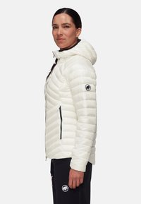 Giacca imbottita bianca con cappuccio, caratterizzata da cuciture diagonali, tasche con zip e un logo sulla manica. Tessuto dalla superficie liscia.