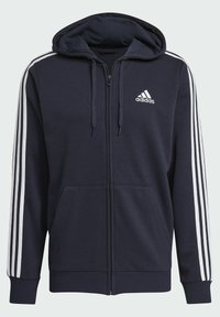 Sudadera con cremallera de color azul marino, hecha de una tela suave, con una gran capucha, bolsillos frontales, logo de Adidas en blanco y acentos de tres rayas blancas en las mangas.