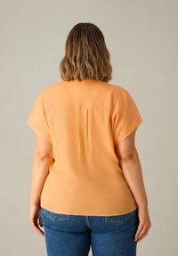 Blusa a maniche corte in morbido tessuto arancione, con una vestibilità rilassata, dettaglio a piega sul retro e una superficie liscia. Abbinata a jeans in denim blu.