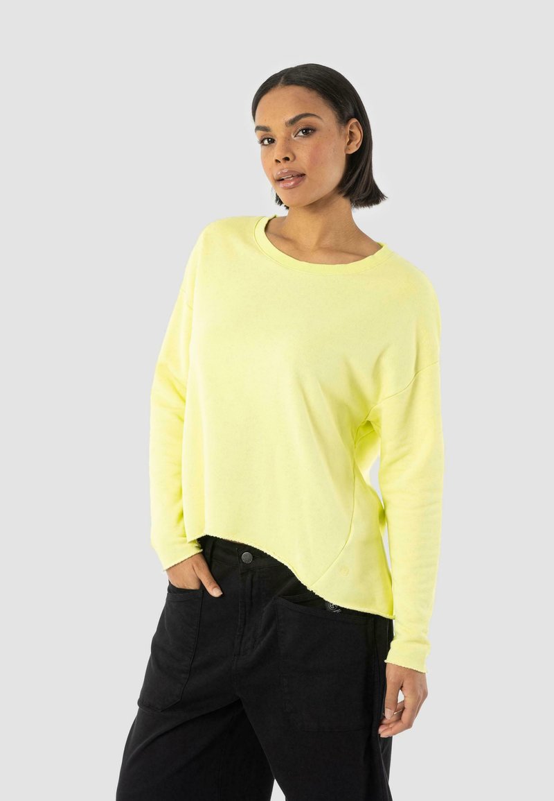 Světle žlutý crew-neck svetr s volným střihem a asymetrickým lemem. Černé kalhoty se bočními kapsami doplňují celý outfit.