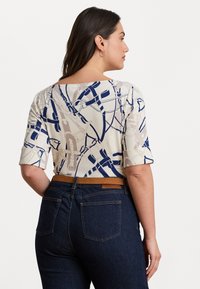 Kratka majica s kratkimi rokavi v kremni barvi z modrim abstraktnim vzorcem, z bateau izrezom. V kombinaciji z temno jeans hlače srednjega pasu in rjavim pasom.