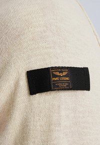 Beige gebreide trui met een zwart stoffen label met oranje tekst waarop staat "PME LEGEND" en "GETEST BIJ HOGE HOOGTE."