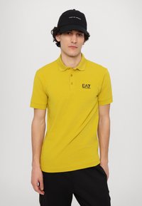 Een gele polo met korte mouwen en een kraag, voorzien van een zwart EA7-logo op de linkerkant van de borst. De achtergrond is eenvoudig wit.