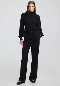 Maglione nero a coste con collo alto e maniche a sbuffo trasparenti, abbinato a pantaloni neri a vita alta con cintura di perline e finitura liscia.