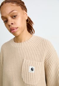 Maglione lavorato a maglia beige con scollatura rotonda, dotato di tasca frontale e patch con logo. La texture è a coste e confortevole.