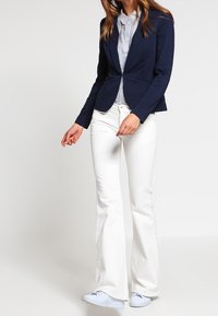 Blazer bleu marine à la coupe ajustée, associé à une chemise rayée bleu clair et à un jean évasé blanc. Des chaussures bleu clair complètent la tenue.
