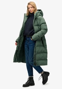 Langer, gepolsterter grüner Parka mit Kapuze, ausgestattet mit einem Reißverschluss und Knopfakzenten. Getragen über einem marineblauen Pullover und blauen Jeans mit schwarzen Schnürstiefeln.