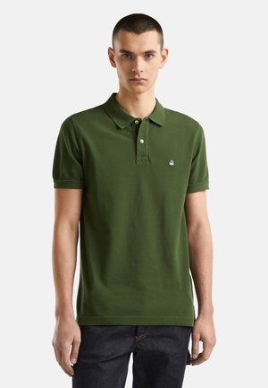 Młody mężczyzna z krótkimi włosami, ubrany w zieloną koszulkę polo z krótkim rękawem i ciemne dżinsy, stojący na tle jasno jednolitego tła.