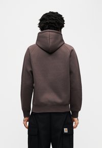 Καφέ φούτερ με κουκούλα και κορδόνι, με ριγέ μανσέτες και τελείωμα. Το ύφασμα έχει λεία υφή και διαθέτει μινιμαλιστικό σχέδιο χωρίς ορατά λογότυπα.