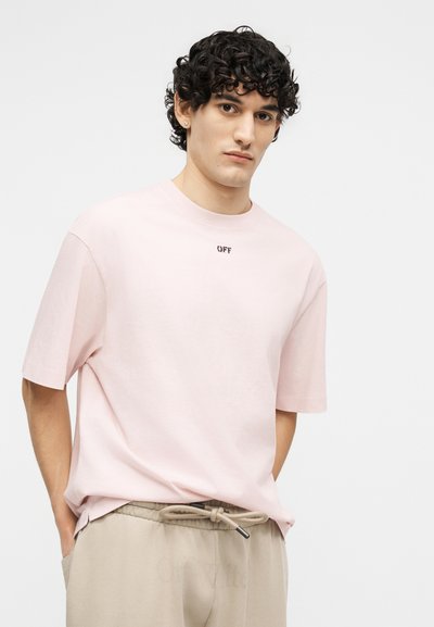 Άνδρας με σγουρά σκουρόχρωμα μαλλιά που φοράει ανοιχτό ροζ φαρδιά μπλούζα με το κείμενο "OFF" και μπεζ παντελόνι με κορδόνι στη μέση, τα χέρια στις τσέπες, ουδέτερο φόντο.