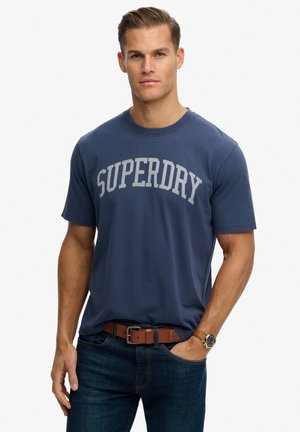 Man standing with one hand in pocket wearing blue "SUPERDRY" t-shirt, dark jeans, brown belt, and watch on left wrist.
