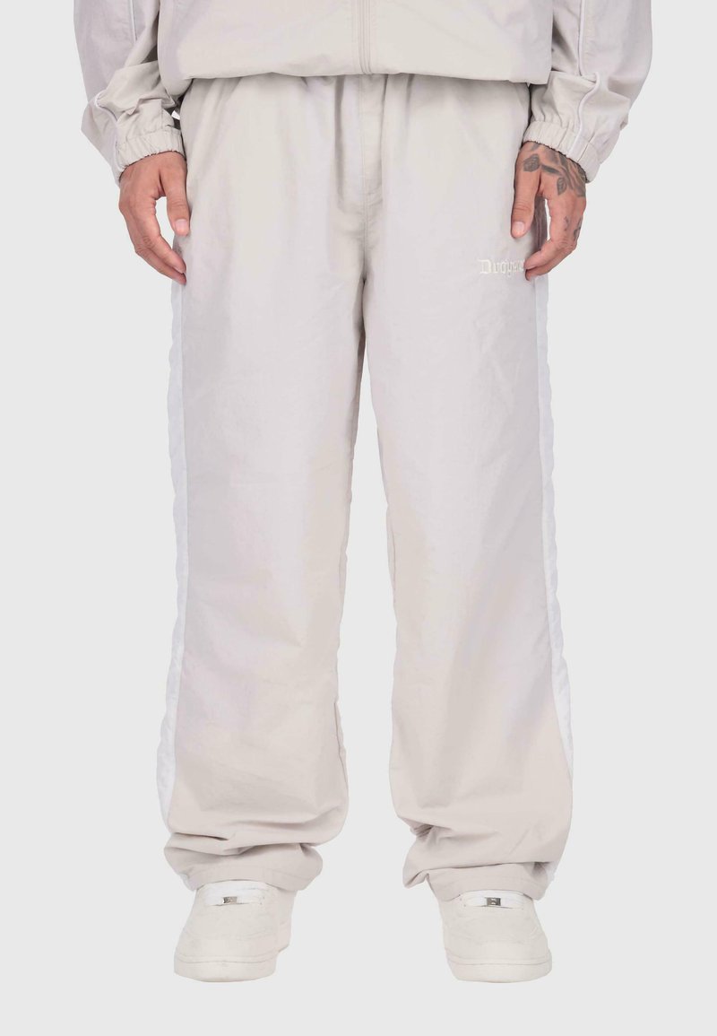 Persona ar gaiši bēšiem sporta biksēm ar baltām sānu līnijām, apvienotiem ar saskaņojošiem sporta apaviem un gaiši bēšu jaku, stāv taisni.