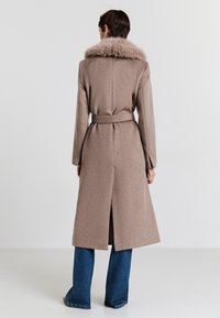 Lungo cappotto marrone con colletto in pelliccia sintetica, vita con cintura e spacco sul retro, abbinato a jeans blu a zampa d'elefante. Texture liscia.