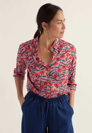 Sötét hajú nő lófarokkal, rózsaszín virágmintás gombos ingben és kék nadrágban, kezét zsebében tartva, jobbra néz.