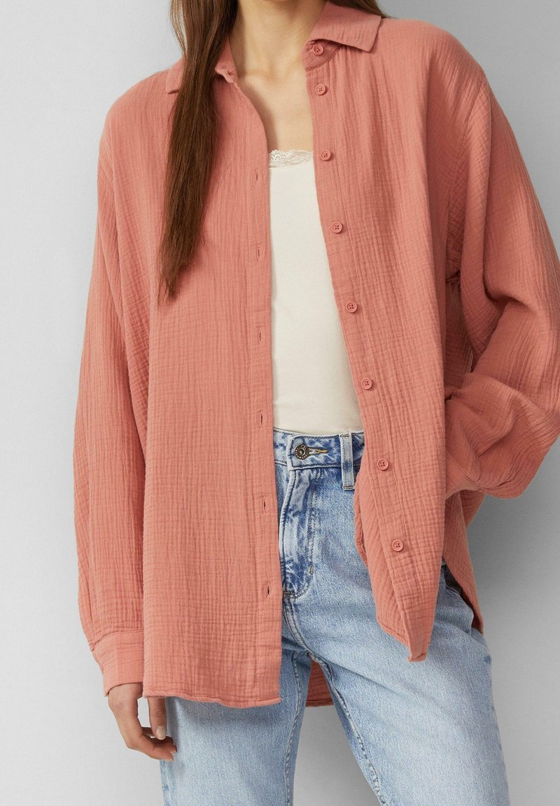 Donna che indossa una camicia button-up rosa polverosa e larga sopra una canotta bianca con pizzo e jeans a vita alta di un blu chiaro.