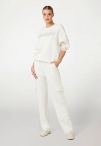 Hanorac alb cu logo-ul „GUESS” argintiu, asortat cu pantaloni cargo albi cu buzunare. Textură netedă și croială lejeră.