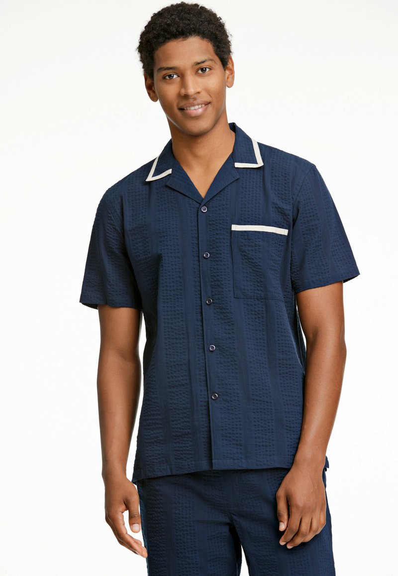 Camisa de pijama de manga corta en color navy con un patrón texturizado, ribete blanco en el cuello y el bolsillo, y cierre frontal con botones. Presenta un bolsillo en el pecho.