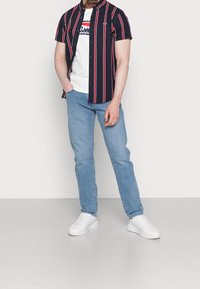 Kortærmet, marineblå stribet skjorte med røde og hvide striber, parret med lyseblå jeans og hvide sneakers, stående på en ensfarvet baggrund.