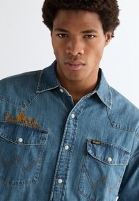 Νέος άνδρας που φοράει πουκάμισο από τζιν με κουμπιά, διακοσμημένο με κεντημένο σχέδιο καουμπόη και κάκτου στις τσέπες του στήθους, κοιτάζοντας μπροστά σε απλό φόντο.