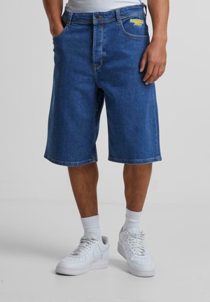 Denim šorti vidēji zilā krāsā ar brīvu piegriezumu, pieci kabatas un logotipa izšuvums priekšpusē, pārī ar baltām sporta kurpēm.