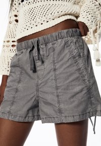 Primer plano de una persona que lleva pantalones cortos grises con cordón y un suéter de crochet crema con largas borlas en las mangas.