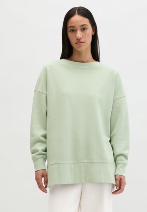 Młoda kobieta o długich ciemnych włosach, ubrana w luźną, jasnozieloną bluzę i białe spodnie, stojąca na tle jednolitego, białego tła.