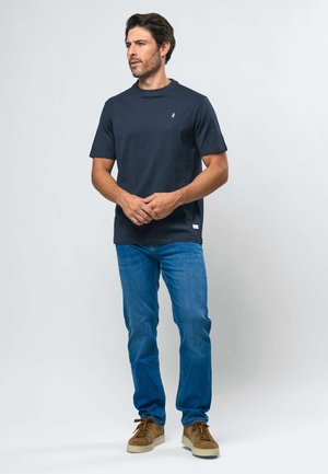 Mand iført mørkeblå kortærmet skjorte, blå jeans og brune sko, stående med hænderne foldet, kiggende til siden mod en ensfarvet baggrund.