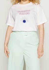 Camiseta de algodón blanca con "VISIÓN CÓSMICA" en fuente rosa y un gráfico de un ojo con un iris azul. Combinada con pantalones de pierna ancha de color menta claro.