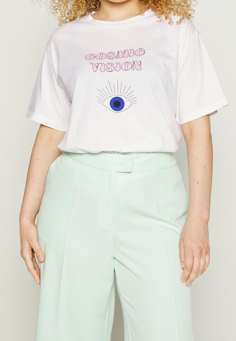 Camiseta de algodón blanca con "VISIÓN CÓSMICA" en fuente rosa y un gráfico de un ojo con un iris azul. Combinada con pantalones de pierna ancha de color menta claro.