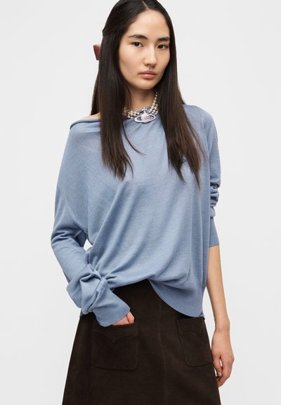 Ανοιχτό μπλε φαρδύ πουλόβερ με ριμπ μανσέτες και ασύμμετρο τελείωμα, συνδυασμένο με μια σκουρόκαφέ φούστα από σουέντ με τσέπες.