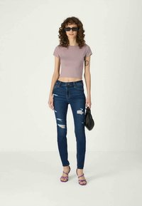 Gaiši purpura krūšturis, pieguļošas zila džinsas bikses ar nolietotiem apgabaliem, purpura sandaļi un melna somiņa. Kļūdainas mati, valkā tumšas saulesbrilles.