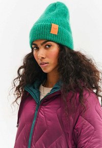 Zelená pletená čepice s žebrovanou texturou a malou oranžovou štítkem. Nošena s vínově červenou péřovou bundou s tyrkysovým límcem a detailním prošitím.