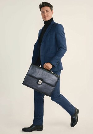 Granatowy garnitur w kratę o slim fit, zestawiony z czarnym golfem i gładkimi czarnymi skórzanymi butami. Niesie czarną skórzaną teczkę z metalowymi akcentami.