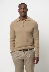 Beige gerippter Strickpullover mit Kragen und drei Knöpfen, kombiniert mit hellbraunen gemusterten Hosen und einem braunen Leder-Gürtel.