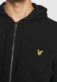 Černá mikina s kapucí na zip vyrobená z měkké tkaniny, s žlutým vyšívaným logem na levé straně hrudi a nastavitelnými šňůrkami na kapuci.