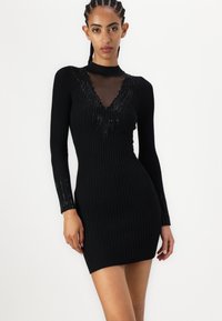 Abito mini nero a costine con maniche lunghe, caratterizzato da un colletto trasparente e dettagli decorativi, che creano una silhouette aderente.