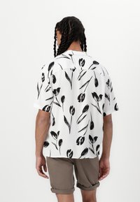Camisa de manga corta en blanco con un patrón floral negro, que cuenta con un corte relajado, textura suave y un dobladillo recto en la parte trasera.