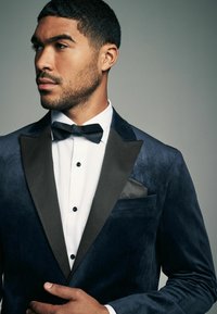 Marineblaues Samt-Tuxedojackett mit schwarzen Satin-Revers, strukturiertem Design, weißem Hemd, Fliege und schwarzem Einstecktuch mit Muster.