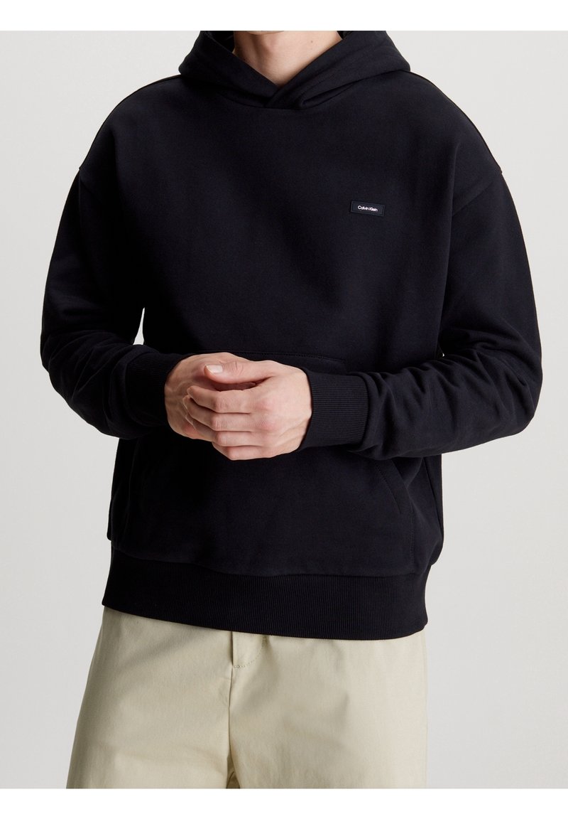 Persona con una sudadera negra de Calvin Klein, con las manos entrelazadas al frente, combinada con pantalones beige, contra un fondo simple.