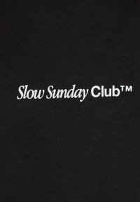 Musta kangas, jossa on valkoista tekstiä, joka lukee "Slow Sunday Club™." Pintastruktuuri on sileä ja hienovarainen.