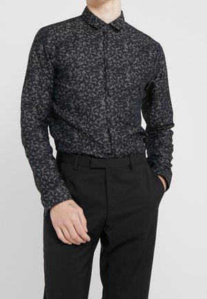 Mann trägt ein slim-fit schwarzes Hemd mit grauem abstraktem Muster, in schwarze Anzugshosen gesteckt, Hände locker an den Seiten.