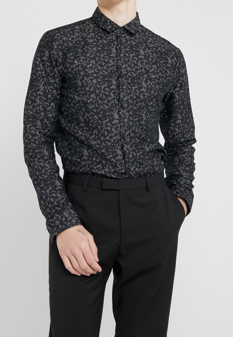 Homme portant une chemise noire slim-fit avec un motif abstrait gris rentrée dans un pantalon habillé noir, les mains détendues le long du corps.