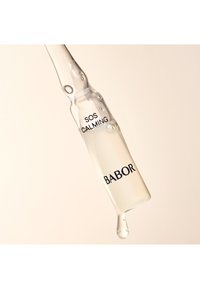 Klar glasampull som innehåller en ljusgul vätska märkt "SOS CALMING" och "BABOR" i svart text, med en droppe som bildas i spetsen.