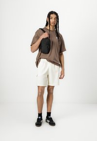 Brūns pārmērīgs T-krekls, baltas šortes, melna plecu soma. Melni sporta apavi ar baltām akcentiem. Vienkrāsains fons, stāvoša poza.