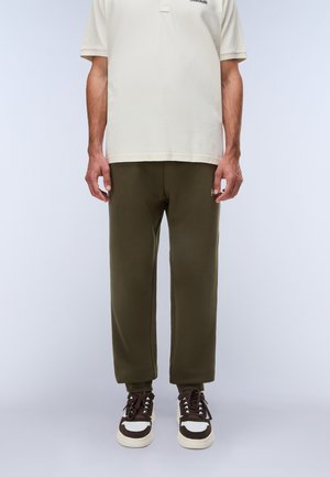 Mand iført olivengrønne joggingbukser, brune og hvide sneakers og en cremefarvet poloshirt med korte ærmer, står foran en ensfarvet baggrund.