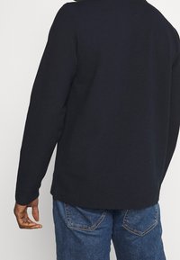 Dunkelblauer Strickpullover mit langen Ärmeln, gerippter Textur und geradem Saum. Kombiniert mit blauen Jeans mit Gesäßtaschen.